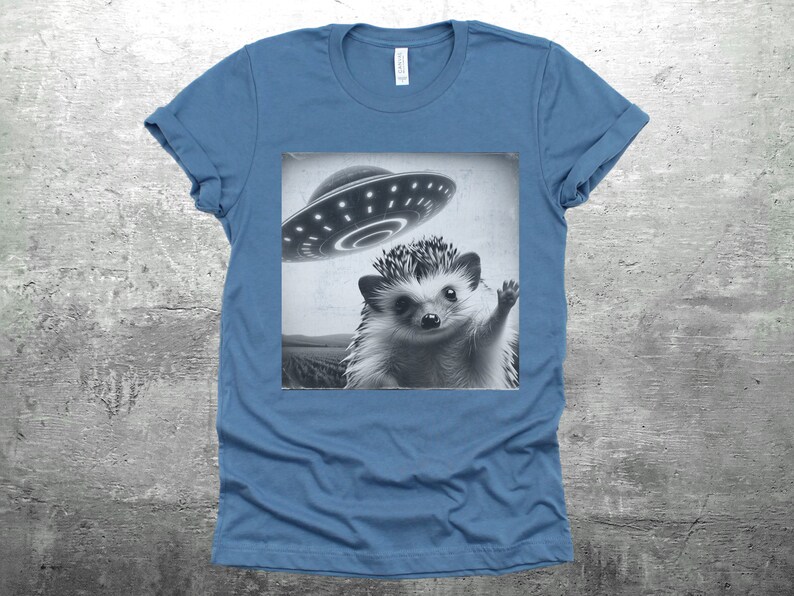 May include: A blue t-shirt with a black and white image of a hedgehog waving at a flying saucer.