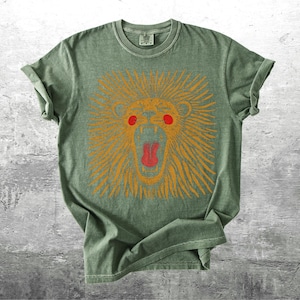 May include: A sage green t-shirt with a graphic of a lion's head. The lion is depicted with orange fur, red eyes, and a red open mouth. The shirt has short sleeves and a crew neck.