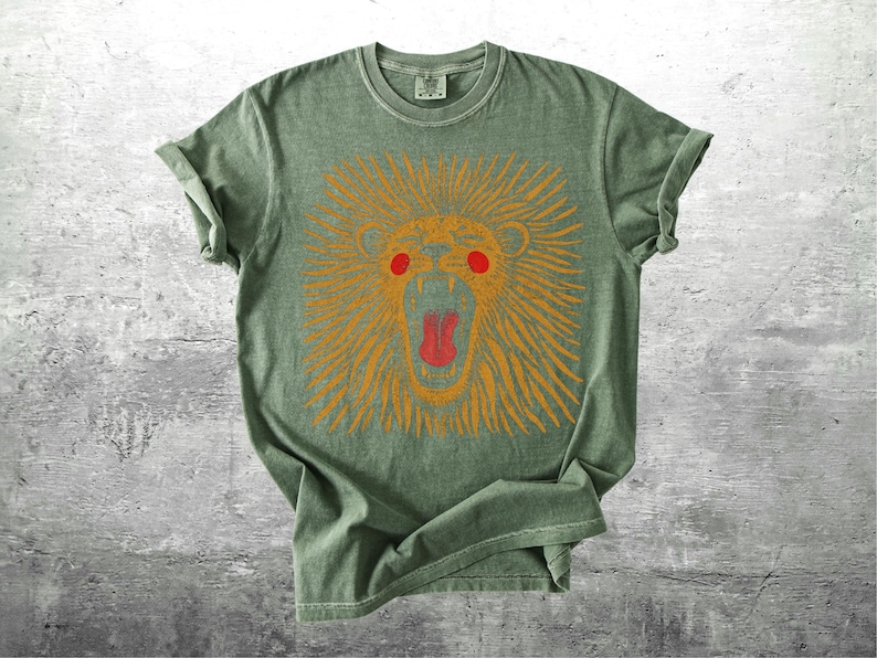 May include: A sage green t-shirt with a graphic of a lion's head. The lion is depicted with orange fur, red eyes, and a red open mouth. The shirt has short sleeves and a crew neck.