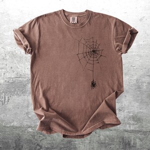 May include: Brown t-shirt featuring a black spider web design with a spider hanging from the web. The shirt has a crew neck and short sleeves. The fabric has a slightly faded appearance.