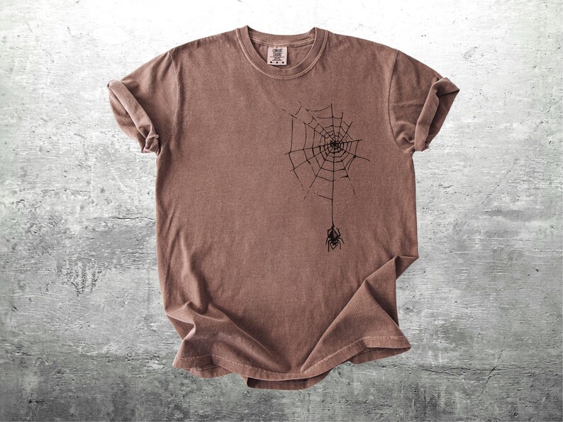 May include: Brown t-shirt featuring a black spider web design with a spider hanging from the web. The shirt has a crew neck and short sleeves. The fabric has a slightly faded appearance.
