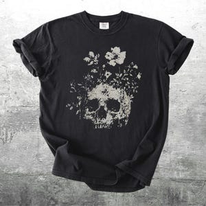 Pu&ograve; includere: T-shirt nero con un design a teschio invecchiato adornato da elementi floreali grigi e bianchi. Il design &egrave; centrato, offrendo un'estetica gotica e botanica.