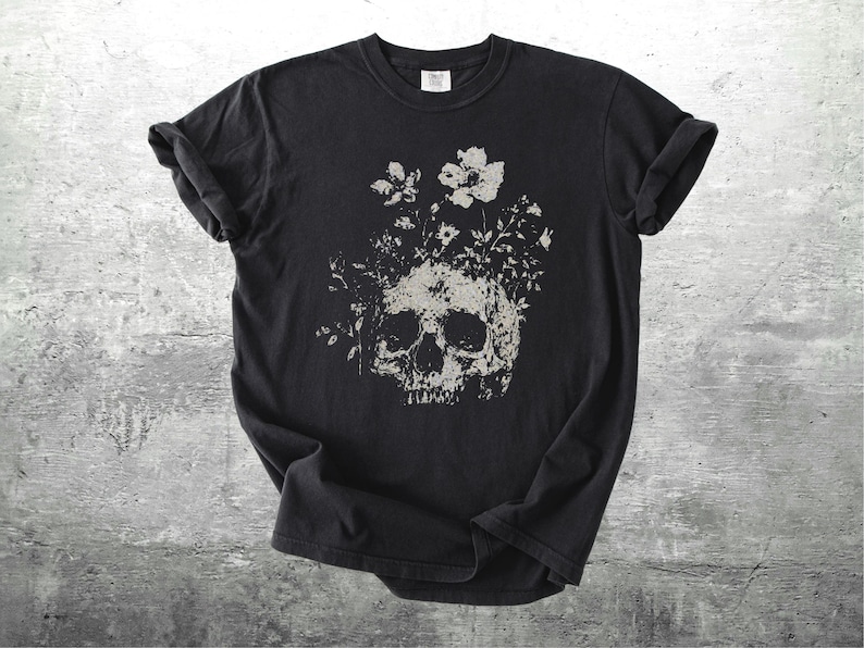 Pu&ograve; includere: T-shirt nero con un design a teschio invecchiato adornato da elementi floreali grigi e bianchi. Il design &egrave; centrato, offrendo un'estetica gotica e botanica.