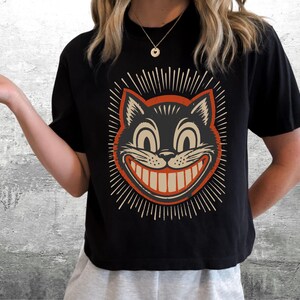 May include: A black short-sleeved t-shirt featuring a graphic print of a grinning cat face with wide eyes and a large toothy smile. The cat illustration is framed by a circular orange border and radiating lines in a light cream color.
