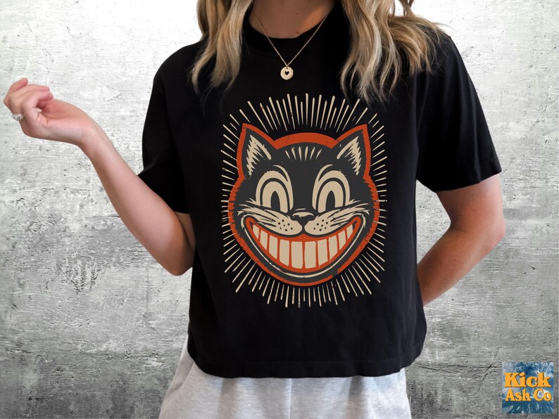 May include: A black short-sleeved t-shirt featuring a graphic print of a grinning cat face with wide eyes and a large toothy smile. The cat illustration is framed by a circular orange border and radiating lines in a light cream color.