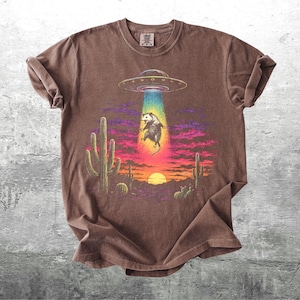 May include: A brown short-sleeve t-shirt featuring a graphic print of a possum being abducted by a flying saucer. The scene includes a desert landscape with cacti, a sunset, and a vibrant rainbow-colored light beam emanating from the UFO.