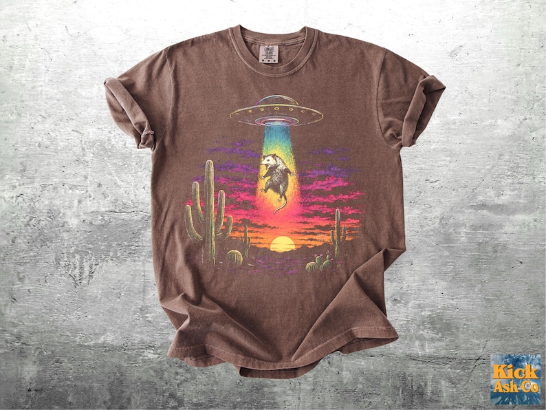 May include: A brown short-sleeve t-shirt featuring a graphic print of a possum being abducted by a flying saucer. The scene includes a desert landscape with cacti, a sunset, and a vibrant rainbow-colored light beam emanating from the UFO.