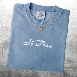 May include: A light blue t-shirt with the text "forever dilly dallying" in white embroidery. The shirt has a distressed look and is made by Comfort Colors.