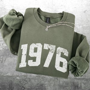 May include: An olive green sweatshirt with the year "1976" printed in distressed white lettering. A silver necklace with a small pendant rests on the folded garment. The sleeves are cuffed.