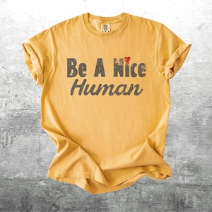 K&ouml;nnte beinhalten: Senfgelbes T-Shirt mit hochgekrempelten &Auml;rmeln. Das Shirt tr&auml;gt den Text "Be A Nice Human" in abgenutzten grauen Buchstaben, mit einem kleinen roten Herz &uuml;ber dem Wort "Nice". Das Shirt ist aus einem weichen Material.
