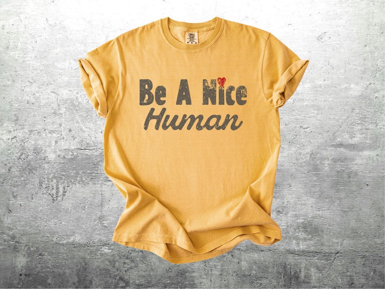 K&ouml;nnte beinhalten: Senfgelbes T-Shirt mit hochgekrempelten &Auml;rmeln. Das Shirt tr&auml;gt den Text "Be A Nice Human" in abgenutzten grauen Buchstaben, mit einem kleinen roten Herz &uuml;ber dem Wort "Nice". Das Shirt ist aus einem weichen Material.