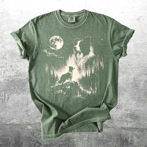 Pu&ograve; includere: T-shirt verde con una grafica di un cane in una foresta sotto la luna piena. Il design &egrave; in tonalit&agrave; di bianco e beige, con la silhouette del cane e i dettagli della foresta. La maglietta ha le maniche arrotolate.