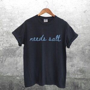 May include: A black t-shirt with the text "needs salt" written in blue cursive.