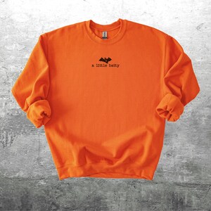 May include: Bright orange sweatshirt with the phrase "a little batty" printed in black, along with a bat graphic. The long-sleeved crew neck shirt is made of a soft material. The shirt is a Halloween-themed apparel item.