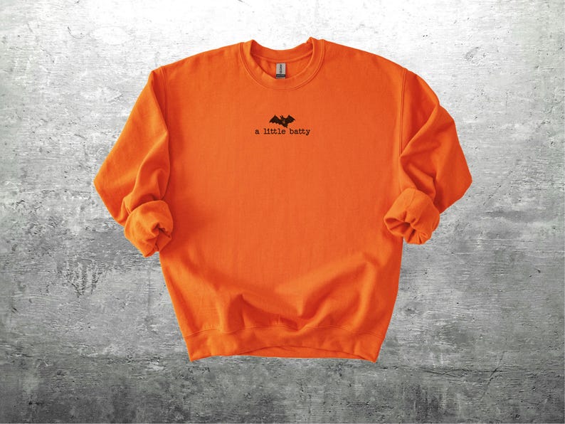 May include: Bright orange sweatshirt with the phrase "a little batty" printed in black, along with a bat graphic. The long-sleeved crew neck shirt is made of a soft material. The shirt is a Halloween-themed apparel item.