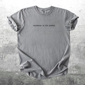 May include: Grey t-shirt featuring the phrase "sunshine is for plants" in a minimalist font. The shirt has rolled-up sleeves and is made of a soft fabric. The background is a textured grey surface.