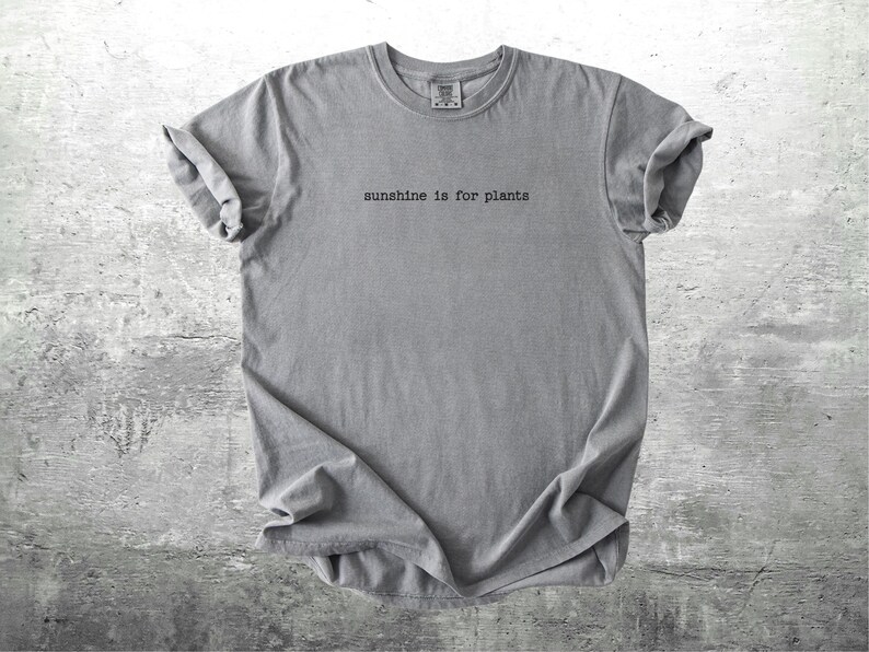 May include: Grey t-shirt featuring the phrase "sunshine is for plants" in a minimalist font. The shirt has rolled-up sleeves and is made of a soft fabric. The background is a textured grey surface.