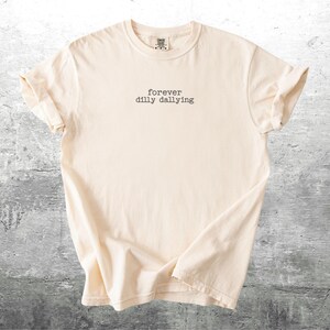 May include: A cream-colored t-shirt with the text "forever dilly dallying" printed in black.