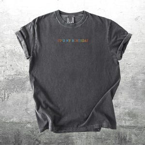 Puede incluir: Una camiseta gris oscuro con el texto "It's My Birthday" bordado en colores arco&iacute;ris.
