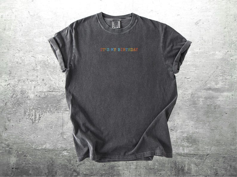 Puede incluir: Una camiseta gris oscuro con el texto "It's My Birthday" bordado en colores arco&iacute;ris.