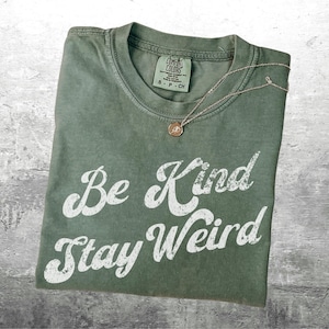May include: An olive green t-shirt with the words "Be Kind Stay Weird" in white script. A delicate gold necklace with a circular pendant rests on the shirt. The shirt is folded and has a tag at the neckline.