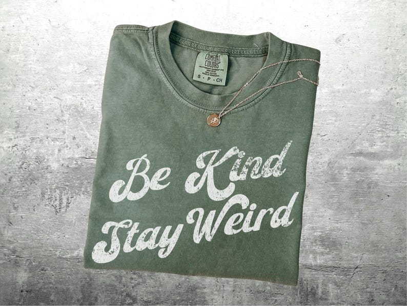 May include: An olive green t-shirt with the words "Be Kind Stay Weird" in white script. A delicate gold necklace with a circular pendant rests on the shirt. The shirt is folded and has a tag at the neckline.