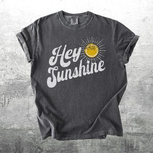 May include: Dark gray t-shirt with rolled-up sleeves. The shirt features the text "Hey Sunshine" in white script, with a yellow sun graphic. The shirt is made of a soft, comfortable material.