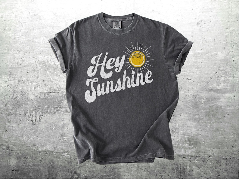 May include: Dark gray t-shirt with rolled-up sleeves. The shirt features the text "Hey Sunshine" in white script, with a yellow sun graphic. The shirt is made of a soft, comfortable material.