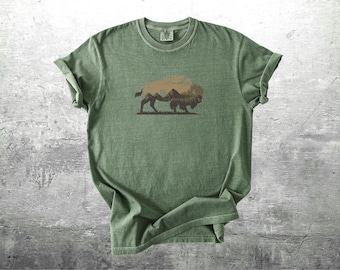 Pode incluir: Camiseta verde com um gráfico de bisão e um design de paisagem de montanha. O bisão é marrom e as montanhas são marrons mais escuras. A camisa é feita de um material macio e tem mangas enroladas.