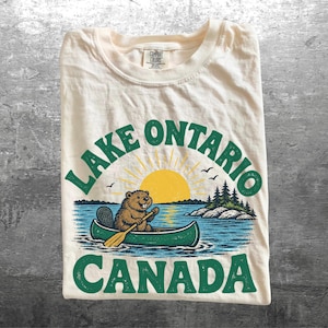 May include: A cream-colored t-shirt featuring a graphic of a brown beaver paddling a green canoe on blue water. Above the beaver is a yellow sun and the text Lake Ontario. Below the canoe is the text Canada. The design includes green pine trees.