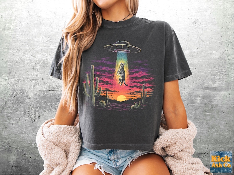 May include: A charcoal gray t-shirt featuring a graphic print of a cat being abducted by a UFO above a desert landscape with cacti and a sunset. The design uses vibrant colors including purple, pink, orange, and teal to depict the scene.