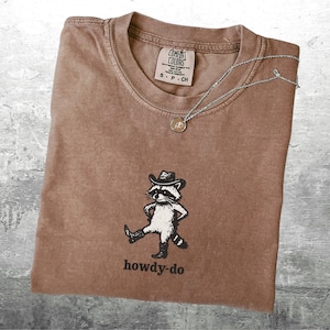 May include: Brown t-shirt with a raccoon wearing a cowboy hat and boots, with the text "howdy-do" below. A silver necklace with a small pendant rests on the shirt. The shirt has a Comfort Colors tag.