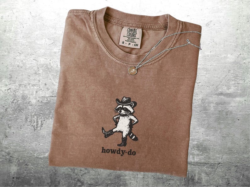 May include: Brown t-shirt with a raccoon wearing a cowboy hat and boots, with the text "howdy-do" below. A silver necklace with a small pendant rests on the shirt. The shirt has a Comfort Colors tag.