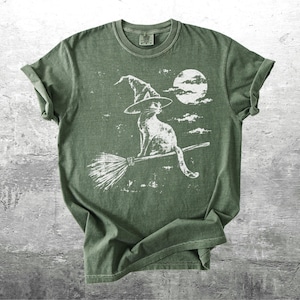 May include: An olive green short-sleeve t-shirt featuring a white graphic print of a cat wearing a witch hat and riding a broomstick in front of a full moon. The design has a distressed, weathered appearance with visible clouds in the background.