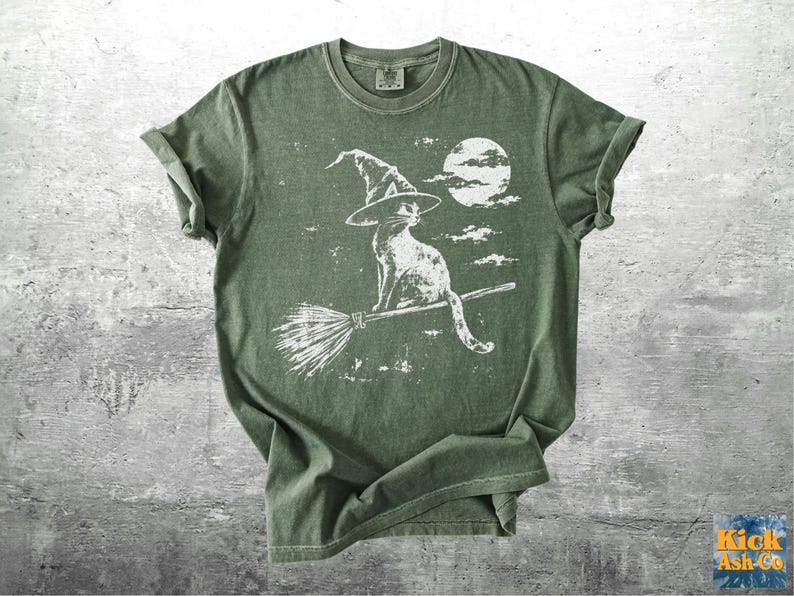 May include: An olive green short-sleeve t-shirt featuring a white graphic print of a cat wearing a witch hat and riding a broomstick in front of a full moon. The design has a distressed, weathered appearance with visible clouds in the background.