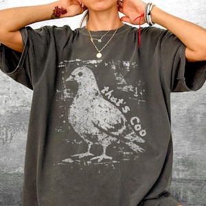 May include: Dark gray t-shirt featuring a white graphic of a pigeon with the text "that's Coo". The shirt has short sleeves and a relaxed fit. The design is distressed, giving it a vintage look.