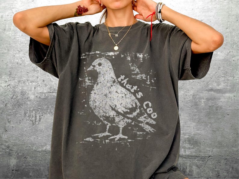 May include: Dark gray t-shirt featuring a white graphic of a pigeon with the text "that's Coo". The shirt has short sleeves and a relaxed fit. The design is distressed, giving it a vintage look.