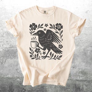 May include: Cream-colored t-shirt featuring a black graphic of a crow holding a steaming coffee cup, surrounded by floral elements. The design is centered on the shirt, with the sleeves rolled up. The shirt is displayed on a textured gray surface.