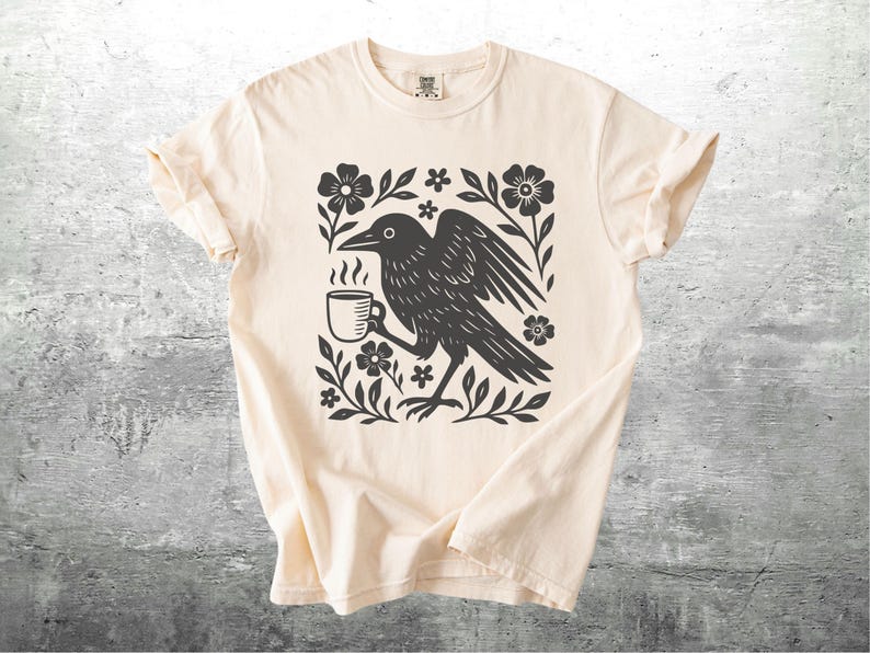 May include: Cream-colored t-shirt featuring a black graphic of a crow holding a steaming coffee cup, surrounded by floral elements. The design is centered on the shirt, with the sleeves rolled up. The shirt is displayed on a textured gray surface.