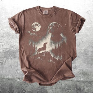 May include: Brown t-shirt featuring a graphic of a dachshund dog silhouetted against a moonlit forest scene. The design includes a full moon and a larger dog image, creating a nature-inspired aesthetic. The shirt has a crew neck and short sleeves.