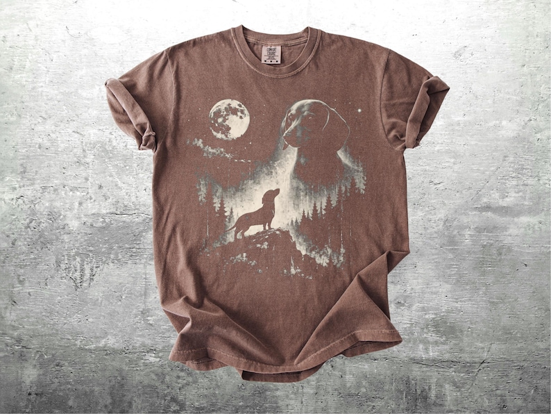 May include: Brown t-shirt featuring a graphic of a dachshund dog silhouetted against a moonlit forest scene. The design includes a full moon and a larger dog image, creating a nature-inspired aesthetic. The shirt has a crew neck and short sleeves.