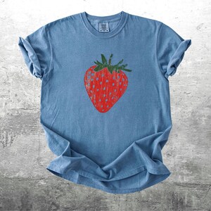 May include: A blue t-shirt featuring a large red strawberry graphic with green leaves. The strawberry design is centered on the front of the shirt. The shirt is made of a soft material and has rolled-up sleeves.
