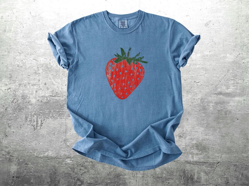 May include: A blue t-shirt featuring a large red strawberry graphic with green leaves. The strawberry design is centered on the front of the shirt. The shirt is made of a soft material and has rolled-up sleeves.