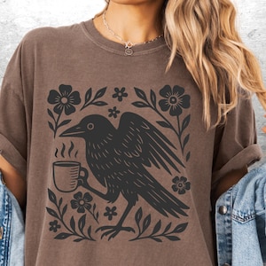 May include: Brown t-shirt featuring a black graphic of a crow holding a coffee cup, surrounded by floral elements. The design is printed on a short-sleeved shirt. The shirt is a neutral color.