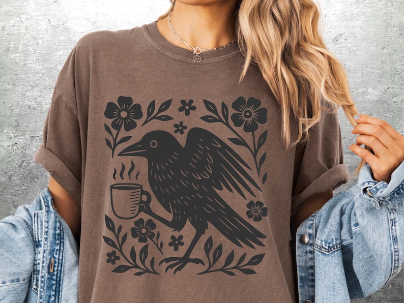 May include: Brown t-shirt featuring a black graphic of a crow holding a coffee cup, surrounded by floral elements. The design is printed on a short-sleeved shirt. The shirt is a neutral color.