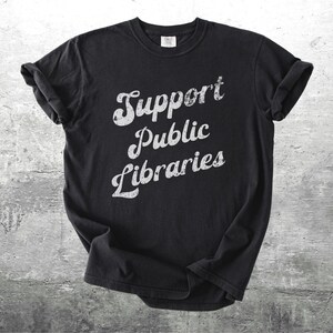 May include: Black t-shirt with rolled-up sleeves. The shirt features the text "Support Public Libraries" in a distressed, white, cursive font. The shirt is displayed on a gray, textured background.