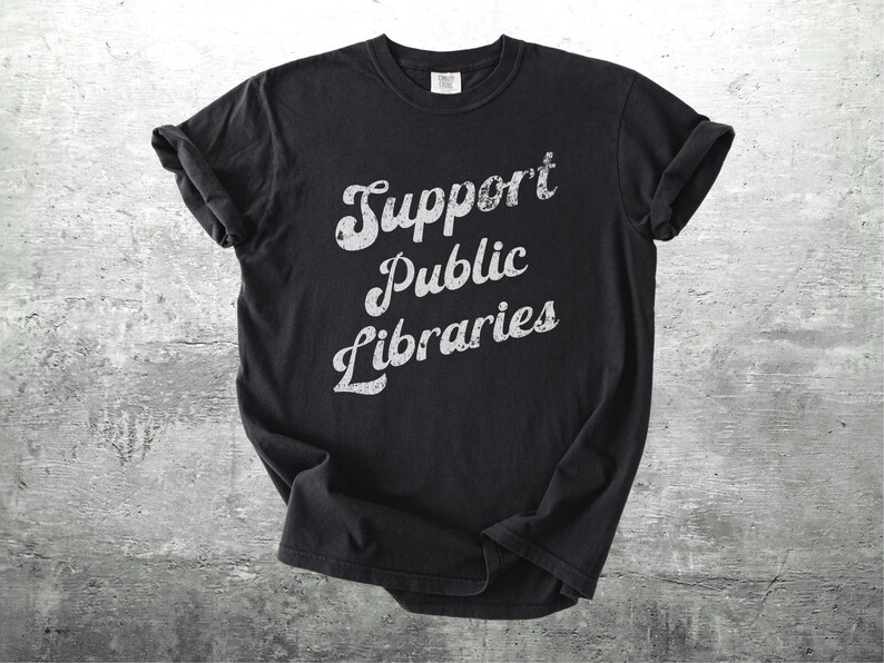 May include: Black t-shirt with rolled-up sleeves. The shirt features the text "Support Public Libraries" in a distressed, white, cursive font. The shirt is displayed on a gray, textured background.