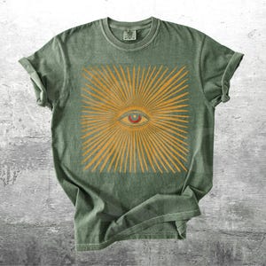 May include: Green t-shirt featuring a square design with an eye at the center, surrounded by radiating lines in a golden-yellow color. The shirt has short sleeves and a crew neck. The shirt is made of a soft, comfortable material.