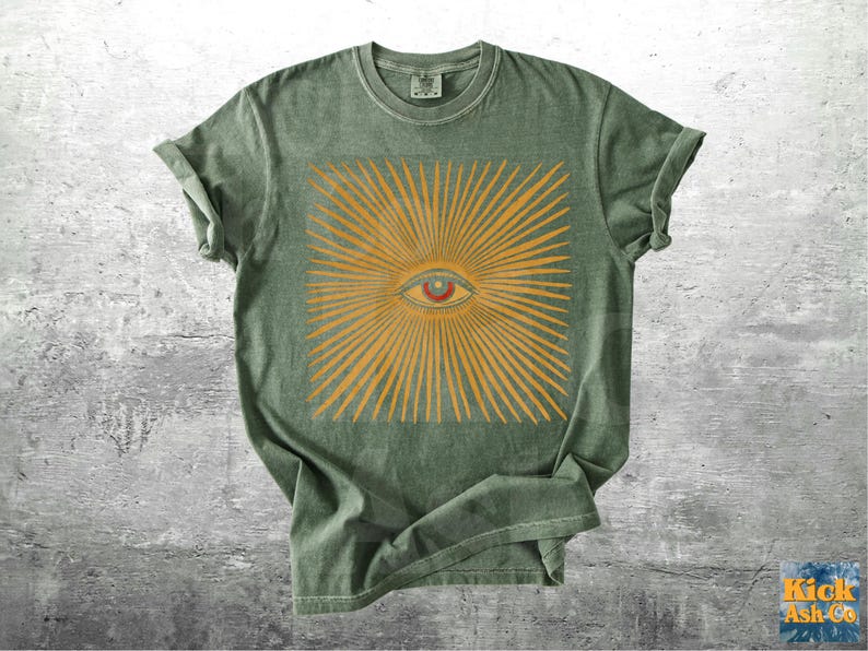 May include: Green t-shirt featuring a square design with an eye at the center, surrounded by radiating lines in a golden-yellow color. The shirt has short sleeves and a crew neck. The shirt is made of a soft, comfortable material.
