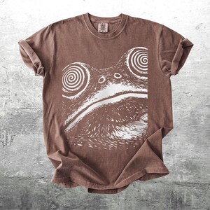 May include: Brown t-shirt featuring a white graphic of a frog's face with hypnotic spiral eyes. The shirt has a crew neck and short sleeves. The fabric appears soft and slightly faded, giving it a vintage look.